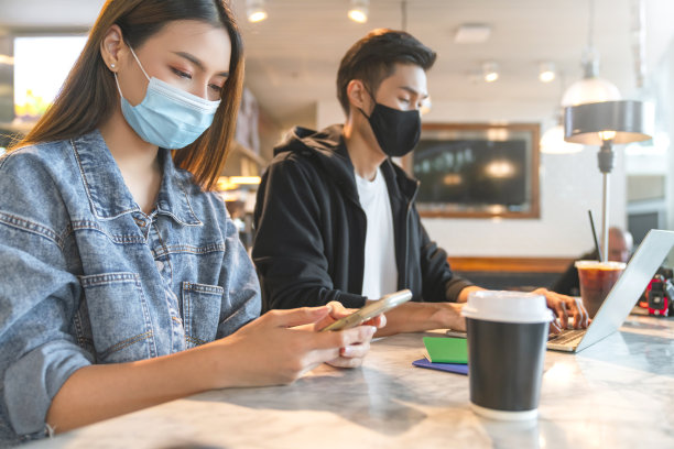 年轻迷人的亚洲女性和男性戴着口罩，保持社交距离，坐在咖啡馆里用笔记本电脑工作素材图片