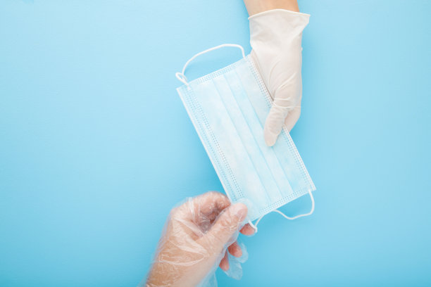 医生用橡胶手套给病人用塑料手套戴口罩。口罩，用以遮盖口鼻。病毒期间的保护。特写镜头。自顶向下的观点。浅粉蓝色的桌子背景。图片下载