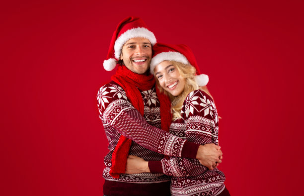 这是圣诞节。一对美丽浪漫的情侣戴着圣诞老人帽，穿着暖和的毛衣，在红色的背景下拥抱在一起图片下载
