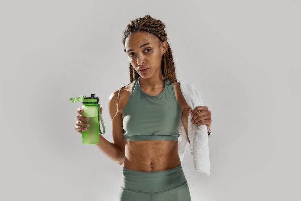 年轻的运动混合种族妇女看镜头，持有蛋白质奶昔鸡尾酒瓶后，强化训练孤立在灰色背景图片下载