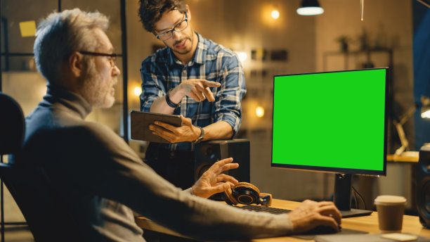 傍晚创意中年男子在桌面电脑绿色模拟屏幕上工作，与使用平板电脑的年轻专家交谈。晚上在大城市的时髦办公室图片下载