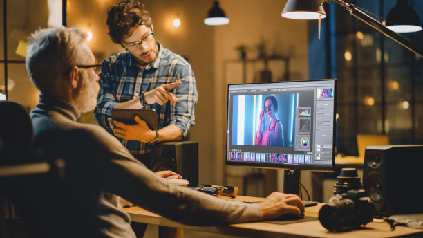 傍晚创意中年照片编辑在台式电脑上使用图像编辑软件，与使用平板电脑的年轻专家交谈。晚上时尚的办公室图片下载