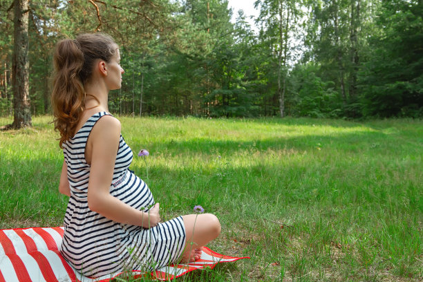 美丽的孕妇坐在草地上的地毯上，闭上眼睛以瑜伽莲花的姿势冥想，把你的腹部放在自然的背景上。图片下载