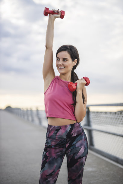 年轻的运动女性，穿着粉红色的运动服，在户外用红色的砝码锻炼图片下载