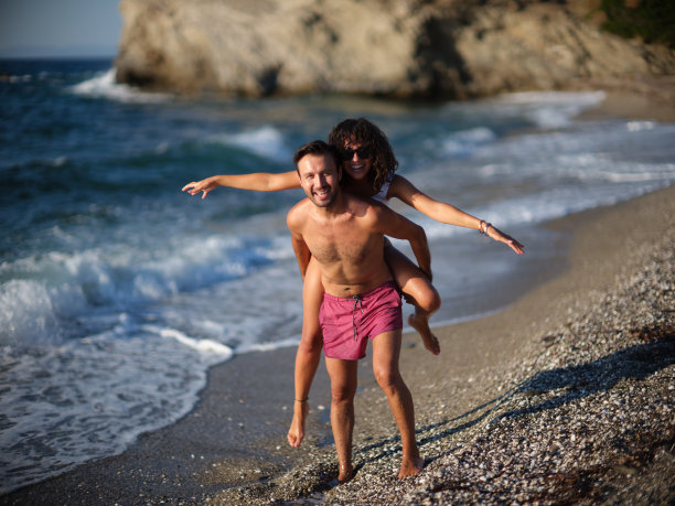 快乐的年轻夫妇-男人在海滩上背着女人图片下载