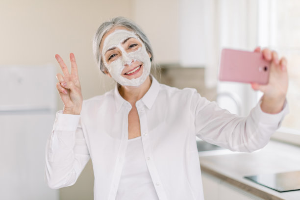 这是一幅欢快迷人的微笑老妇人的肖像，她留着长长的灰色头发，带着泥面膜，在家里厨房对着镜头摆姿势，拿着智能手机，打着胜利的手势图片下载