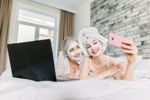 两位微笑的女性，老年母亲和年幼的女儿，在智能手机上做可爱的自拍照片，躺在床上，带着笔记本电脑，脸上戴着面膜。护肤概念图片下载