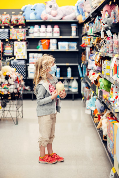 一个新的正常。白种金发女孩戴着卫生面罩在玩具店购物。戴防护口罩的儿童。隔离期间的安全、健康保护。图片下载