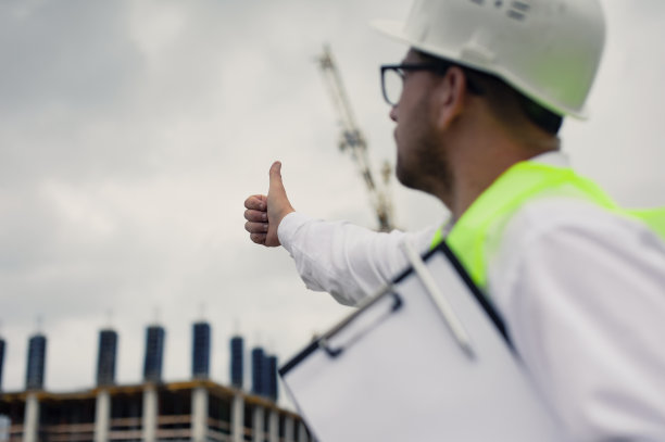 酋长穿着背心，戴着建筑头盔，手里拿着文件，背景是塔吊和布满云彩的天空。住宅建设检查。图片下载