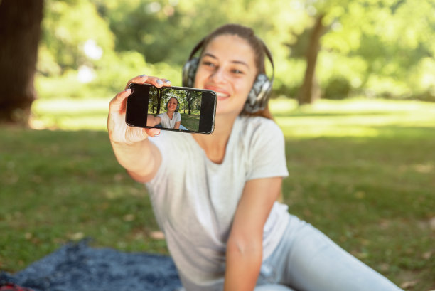 年轻快乐的女孩戴着耳机坐在公园里用智能手机自拍图片下载