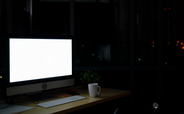 晚上由电脑屏幕照明的房间，没有人，晚上在办公室的一个空的工作场所，从窗户可以看到城市的景色图片下载