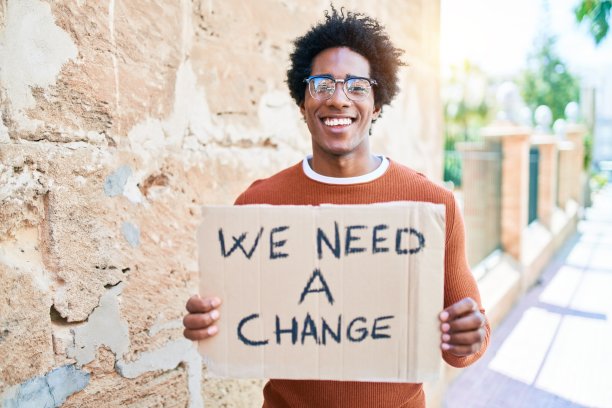 年轻英俊的非裔美国人微笑着请求改变。站在脸上微笑，我们需要一个改变横幅纸板在城镇街道。图片下载