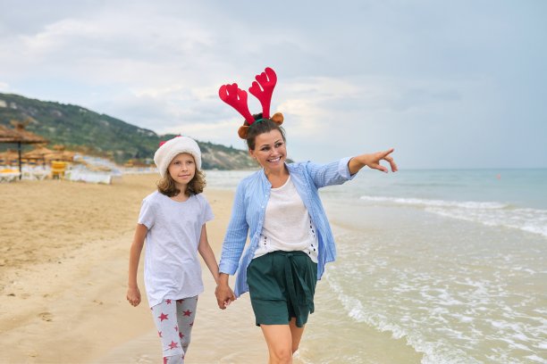 快乐的妈妈和女儿孩子在圣诞老人的帽子沿着海滩散步图片下载