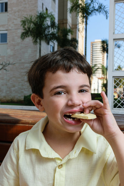 7岁的巴西孩子，在一个阳光明媚的下午，看着镜头，拿着一块白巧克力，咬着(垂直照片)。图片下载
