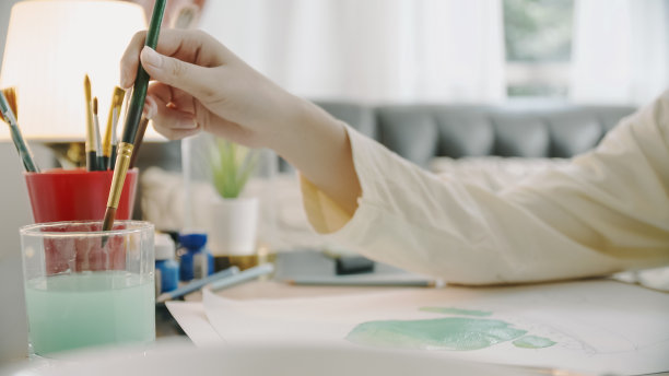 亚洲少女手绘和绘画她的艺术在客厅。她用笔记本电脑和朋友视频通话，同时用水彩和画笔在白纸上创作艺术项目，形成她的想象。家，艺术和手工艺概念。图片下载