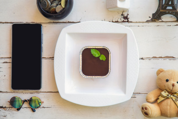 俯视图巧克力软糖蛋糕与薄荷在一个白色的盘子，在一个白色的木制桌子上有一个手机和装饰。图片下载