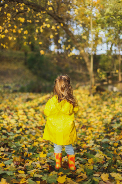 小女孩穿着时髦的黄色雨衣，牛仔裤和橡胶靴站在秋天的森林或公园户外吃纸杯蛋糕。枫叶落在地上。概念秋天家庭照片拍摄图片下载