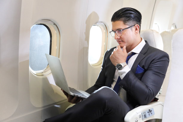 白人商务旅行穿着正式使用笔记本电脑，而舒适地坐在飞机的商务舱窗口座位图片下载