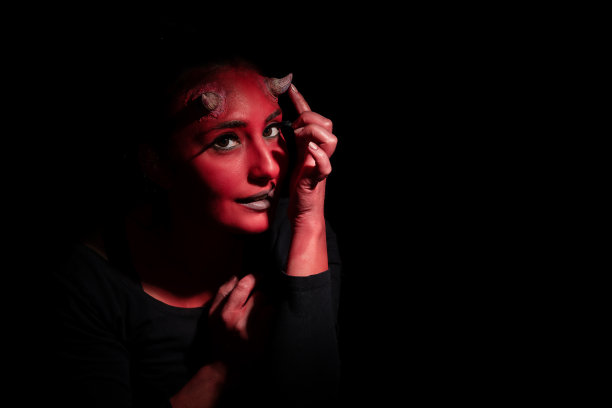 万圣节的概念。妖女大头照，红脸妆，小角，穿着黑色衣服，想让自己看起来毛骨悚然，在黑色背景下摆姿势，有拷贝空间。妖女红脸摸着她的小角图片下载