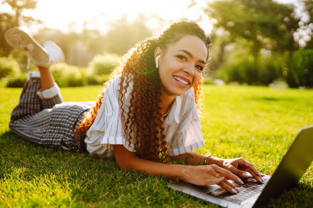 一个漂亮的年轻女子坐在公园的绿草地上，带着笔记本电脑。图片下载
