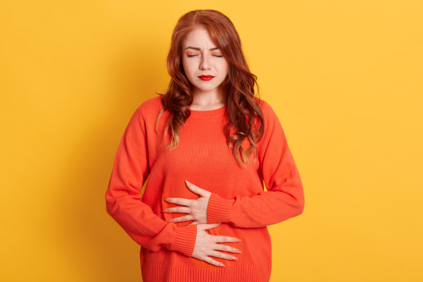 不满意的可爱的年轻女子遭受经期抽筋，保持双手在肚子上，有可怕的疼痛，穿着橙色毛衣，需要医生的帮助。图片下载