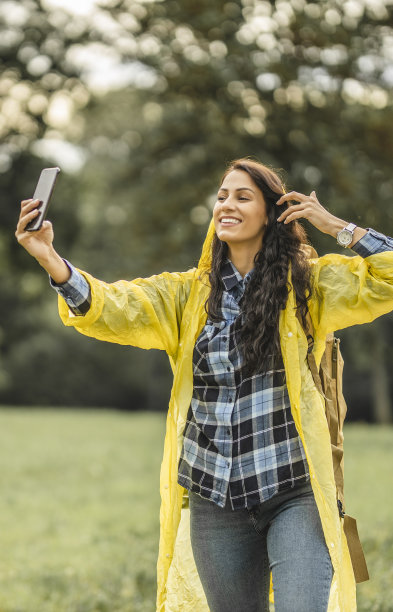 穿着黄色雨衣的年轻女子用智能手机自拍，微笑着享受和休息在自然中图片下载