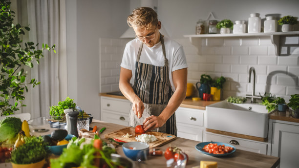 英俊的男人在白衬衫和围裙正在做一个健康的有机沙拉餐在一个现代阳光厨房。戴眼镜的嬉皮士在做饭。自然清洁饮食与健康生活理念。图片下载