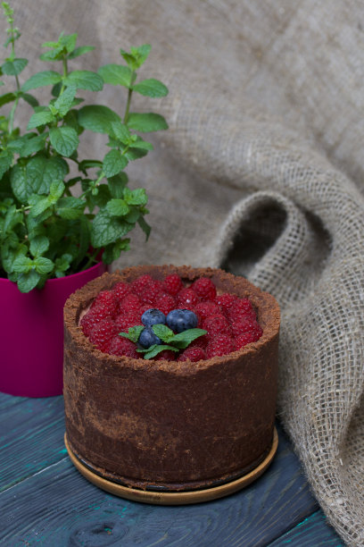 不烤奶油芝士巧克力蛋糕。装饰有覆盆子、蓝莓和薄荷叶。附近有一盆种植着的薄荷。在涂成黑色和绿色的松木板表面。图片下载