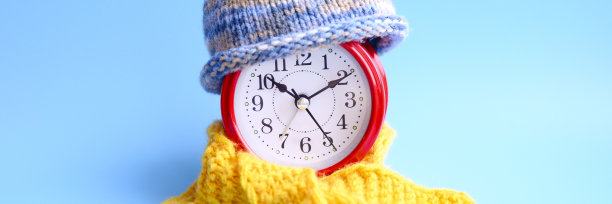 红色圆形闹钟，针织羊毛蓝帽子，黄色围巾，蓝色背景。冬天的时间概念。冬天的季节。舒适和温暖。模拟时间10:10。空间的文本。横幅图片下载