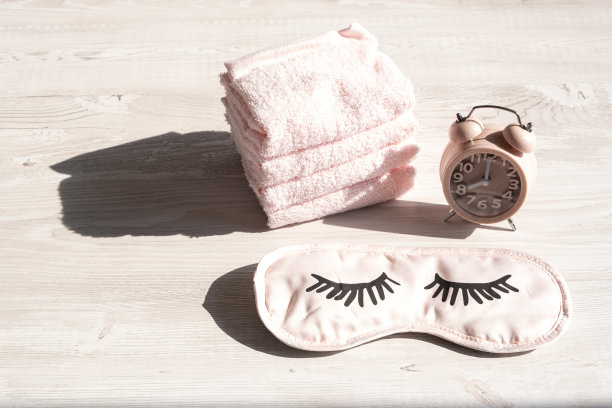 闹钟和睡眠面罩，毛巾放在木制的白色桌子在早上或日落的阳光。早晚的卫生程序，个人护理。本空间图片下载