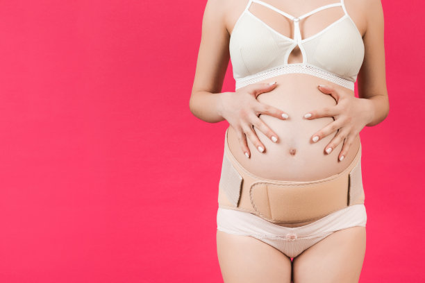 裁剪图像的孕妇在内衣穿着弹性绷带，以防止疼痛在背后的粉红色背景与复制空间。骨科腹部支撑带概念图片下载