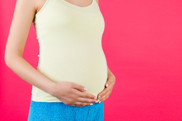 孕妇抱着她的腹部的裁剪图像。妈妈穿着色彩鲜艳的家装，背景是粉红色的。未来的父母期待着她的孩子。本空间图片下载