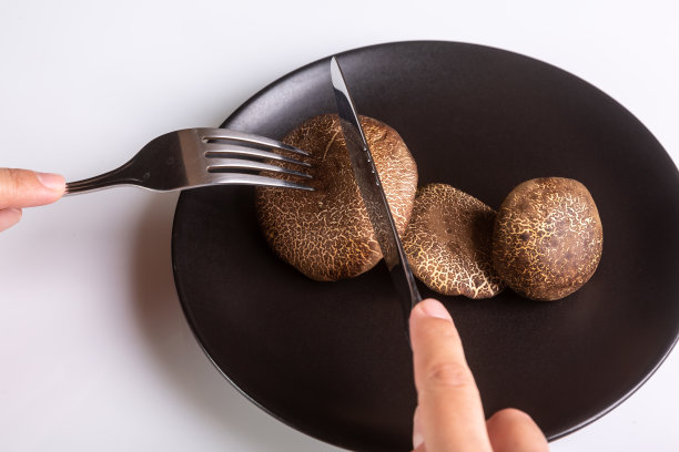 一个人拿着刀叉刀叉，在白色背景的黑色盘子上切着生的泽西牛蘑菇。手在画框里。水平方向。高质量的照片图片下载