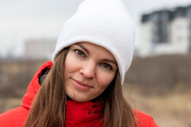 微笑美丽的年轻女子长头发，白色针织帽和一件红色夹克在一个阴天的街道上模糊的背景。女性素颜，自然美。特写视图图片下载