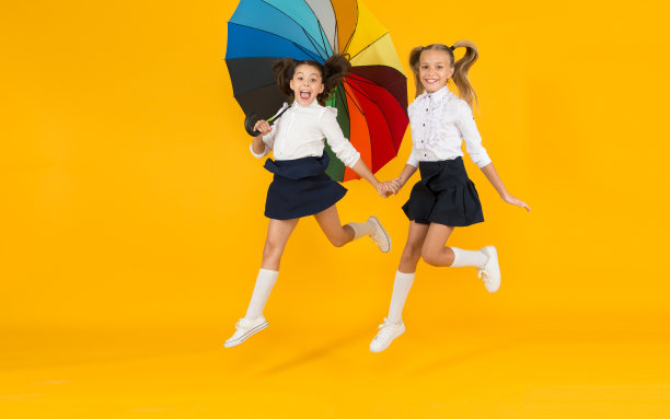 无论什么天气都要回学校。小女孩们在黄色背景的彩色伞下微笑。快乐的小孩子们赶着去学校。享受秋天的学校假期图片下载