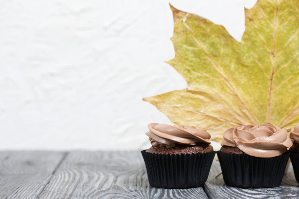 奶油芝士奶油巧克力纸杯蛋糕。附近是干燥的枫叶。在黑色和白色的松木板上。图片下载