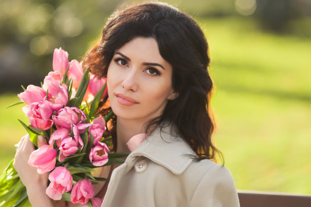 一个春天的一天，年轻严肃的黑发女人穿着时尚休闲装，手里拿着一束粉红色的郁金香，独自坐在公园里等待图片下载