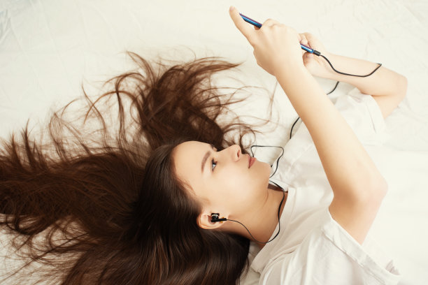 一个年轻的女孩躺在家里的床上，披头散发，看着她的智能手机，听着音乐，为社交网络自拍图片下载