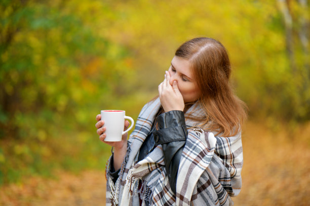流鼻涕的女孩。红头发的女孩捏着鼻子，端着一杯热茶，裹在毯子里以秋天为背景。秋天公园里一个流着鼻涕的生病女人。Gorizontal照片图片下载