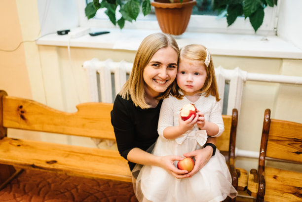 快乐的妈妈和女儿在室内拥抱。一天在幼儿园。孩子和妈妈吃了一个苹果。图片下载