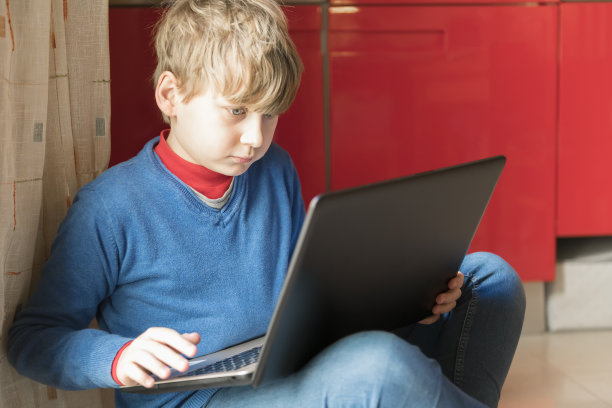 一个金发男孩正坐在房间的地板上，手里拿着笔记本电脑。在家教育和在家在线教育图片下载