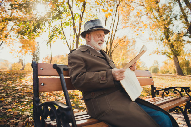 侧面照片的老人有早晨退休，走，坐秋天公园长凳外读镇中心新闻，穿长大衣，帽，帽图片下载