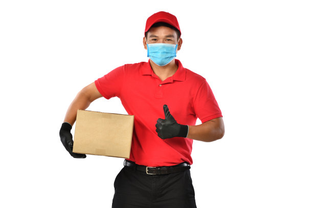 快乐的年轻亚洲快递员身穿红色制服，医疗面罩，防护手套携带一个纸板箱在双手孤立的白色背景。送货员送包裹。安全交付图片下载