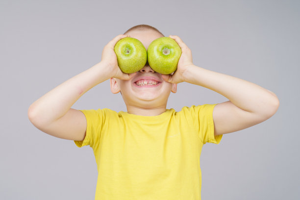 微笑的男孩站在水果，拿着苹果，而不是眼睛。孤立在灰色图片下载