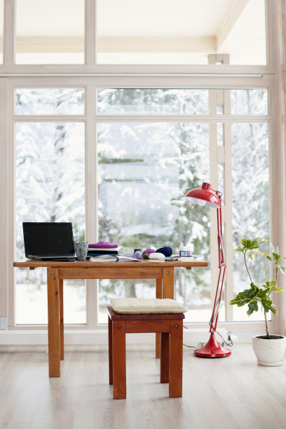 极简风格的工作空间，办公室在一张木桌上，旁边是大落地窗和红色台灯，配有笔记本电脑、纱线和植物。设计师工作的地方。图片下载
