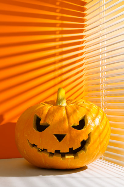 万圣节的节日背景。令人毛骨悚然的万圣节南瓜在一个橙色的背景，被阳光透过百叶窗照亮。图片下载