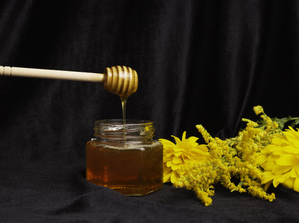 蜂蜜从木铲里滴进玻璃罐里。黄色的花，黑色的背景。有用的农产品。图片下载