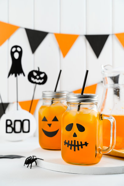 万圣节南瓜冰镇鸡尾酒在玻璃罐装饰可怕的面孔在白色的桌子上。DIY万圣节派对装饰在墙上。图片下载