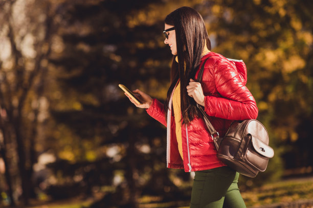 正面的女孩游客的侧面照片，遇见朋友去走秋天公园用智能手机，想找到地方，拿包背包，穿红色外套夹克外套图片下载