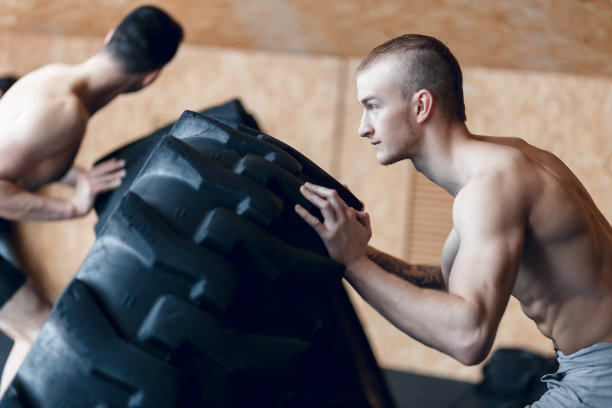 两个健康的男运动员用一个巨大的轮胎在健身房锻炼，旋转和翻转。图片下载